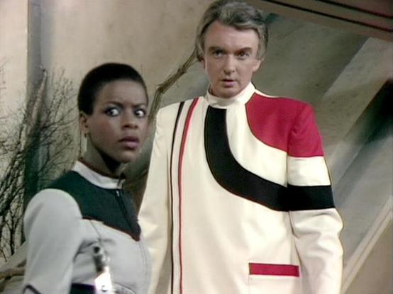 Claude Haiku 4.5 describes the image as: "# Image Description

This scene appears to be from a science fiction television production, likely from the 1980s based on the visual style and costume design. The setting is a stark, minimalist interior space with pale walls and simple furnishings, suggesting a futuristic or institutional environment.

Two actors are featured in the frame. On the left is a woman wearing a dark and white striped uniform that appears to be a space-themed or military-styl…