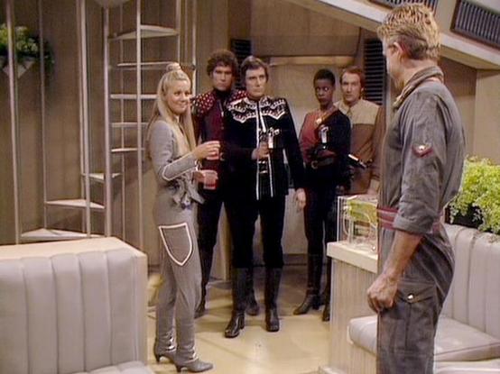 Claude Haiku 4.5 describes the image as: "# Scene from Blake's 7

This image depicts a scene set aboard a spacecraft, featuring a futuristic interior with sleek, minimalist design elements including curved seating, paneled walls, and potted plants. The setting appears to be a command center or common area with a distinctly 1980s science fiction aesthetic.

The scene shows a group of characters in various costumes gathered in conversation. Some wear distinctive black and metallic uniforms typica…