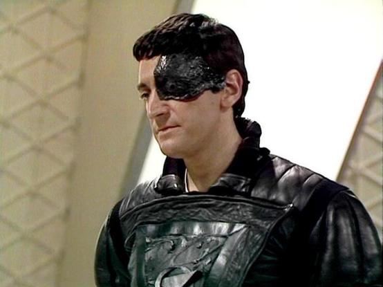 Claude Sonnet 3.7 describes the image as: "The image shows a person in a dark leather-like costume with a distinctive black mask or face covering over one eye. They appear to be on a set with light-colored geometric patterned walls in the background. The costume has a textured, armor-like quality with some embossing or design on the chest piece.

This appears to be a scene from a science fiction or fantasy television production, likely from the late 1970s or 1980s based on the production style …