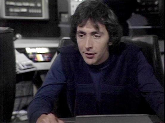 Claude Sonnet 4.5 describes the image as: "This image shows a scene from what appears to be a television production, likely from the 1970s based on the video quality and styling. The setting is an office or control room environment, with electronic equipment visible in the background including what looks like monitors or control panels.

The actor Norman Tipton is seated at a desk or workstation, leaning forward in an engaged posture. He's wearing a dark navy blue zip-up jacket or sweater over …