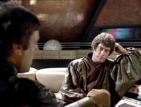 Claude Sonnet 4 describes the image as: "This appears to be a scene from the British science fiction television series "Blake's 7," set aboard what looks like the Liberator spaceship. The image shows two characters in conversation in what appears to be the ship's relaxation area or quarters. One figure is seated on a curved, cream-colored chair or sofa, wearing what appears to be a leather or dark jacket, with curly hair and appearing to be in a contemplative or possibly troubled state with his…