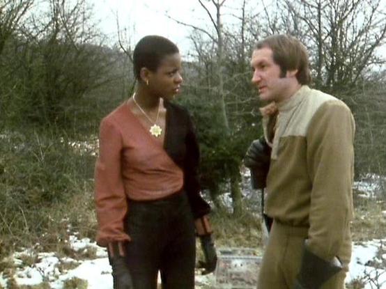 Claude Sonnet 4 describes the image as: "This appears to be a scene from what looks like a British television production, likely from the late 1970s or early 1980s based on the image quality and styling. The setting is outdoors in a wooded, rural area during winter, with patches of snow visible on the ground and bare trees in the background.

The scene shows two people in conversation - one wearing a rust-colored or reddish-brown top with dark pants and a distinctive necklace, and the other in …