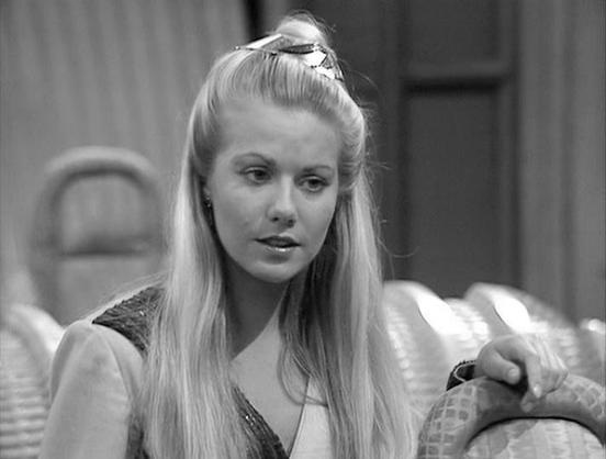 Claude Sonnet 3.7 describes the image as: "This is a black and white image from what appears to be a television production from the 1970s or 1980s. The scene shows a person with long, light-colored hair styled in a half-up fashion with a distinctive top knot or small bun. They're wearing what looks like a decorative or patterned garment with some detailing visible at the neckline. The person appears to be in a conversation or dramatic moment, with their hand resting on what seems to be part of …