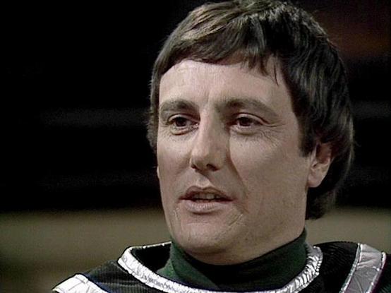 Claude Sonnet 4.5 describes the image as: "This image shows a close-up shot from what appears to be a British science fiction television production, likely from the late 1970s or early 1980s based on the video quality and aesthetic. The frame captures an actor in a distinctive costume featuring a high-collared black and green outfit with striking white and silver decorative elements on the shoulders. The costume design suggests a futuristic or space-opera setting, with its bold geometric patter…