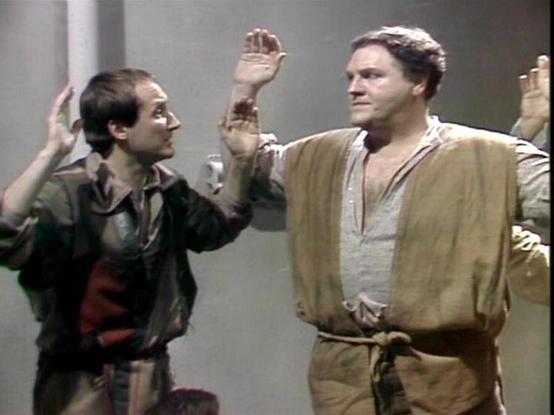 Claude Sonnet 4.5 describes the image as: "This image appears to be from a science fiction television production, likely from the late 1970s or early 1980s based on the video quality and production style. The scene shows several people with their hands raised in what appears to be a gesture of surrender or greeting. 

The setting is minimalist, with plain gray walls creating a stark, institutional atmosphere typical of sci-fi sets from this era. The actors are wearing casual, tunic-style costum…