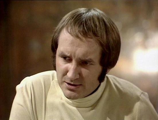 Claude Sonnet 4 describes the image as: "This image shows a man with a distinctive bowl-cut hairstyle wearing a cream or off-white colored garment that appears to be a tunic or simple shirt with a high neckline. The setting appears to be indoors with warm, golden lighting in the background that creates a soft, atmospheric mood. The lighting and overall aesthetic suggest this is from a television production, likely from the 1970s or early 1980s based on the visual quality and styling. The man ha…