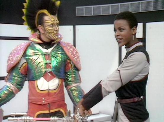 Claude Haiku 4.5 describes the image as: "# Scene Description

This image shows two actors in what appears to be a science fiction setting, likely from a television production. The scene takes place in a modern, minimalist interior with large windows and sleek architectural features visible in the background.

On the left is a character wearing an elaborate costume featuring striking gold and colorful face paint with geometric patterns, along with an ornate outfit composed of a multicolored tun…