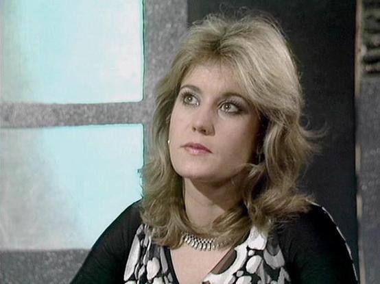 Claude Haiku 4.5 describes the image as: "# Image Description

This appears to be a still from a 1970s television production, capturing a woman with distinctive feathered blonde hair styled in the fashion typical of that era. She wears a black garment with a bold geometric black and white patterned collar or overlay, along with a decorative chain necklace. The lighting and image quality are characteristic of broadcast television from that period.

The setting appears to be an interior space wit…