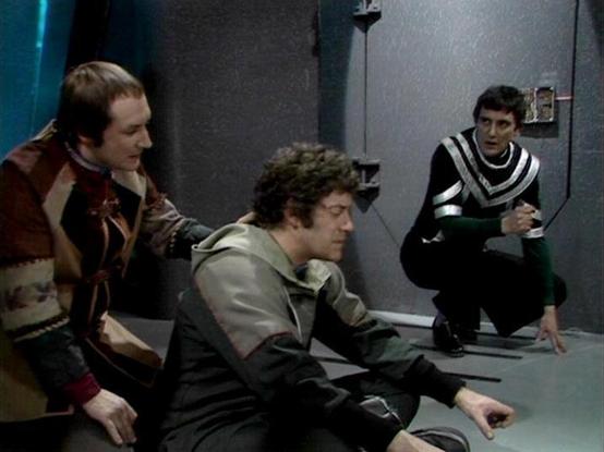 Claude Sonnet 3.7 describes the image as: "The image appears to be a scene from the classic British science fiction series "Blake's 7." It shows three people in a futuristic, sparse gray setting that looks like part of a spacecraft interior. 

The individuals are wearing distinctive costumes typical of the show's aesthetic - one in a brownish patterned jacket, another in a grayish outfit, and the third wearing a black and white striped or chevron-patterned top while crouching down.

This appear…