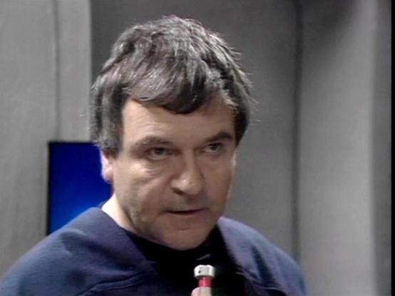 Claude Sonnet 4.5 describes the image as: "This image shows a close-up shot of actor Glyn Owen in what appears to be a scene from a television production, likely from the 1970s based on the video quality and aesthetic. Owen has dark hair with gray at the temples, styled in a period-appropriate cut with a distinctive swept fringe. He's wearing a dark navy or blue crew-neck sweater over what appears to be a black shirt or turtleneck.

The setting appears to be an interior scene with a minimalist,…