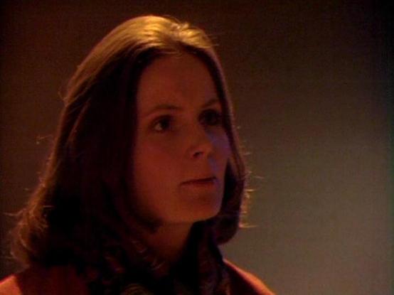 Claude Sonnet 3.7 describes the image as: "The image appears to be a still from what looks like a film or television production with a vintage quality to it. It shows a person with shoulder-length brown hair in a dimly lit setting with warm, reddish lighting. The individual is wearing what appears to be a reddish-brown or orange top or jacket. The scene has a dramatic atmosphere with shadows and low lighting that creates a moody, possibly tense feeling. The cinematography style suggests this mi…