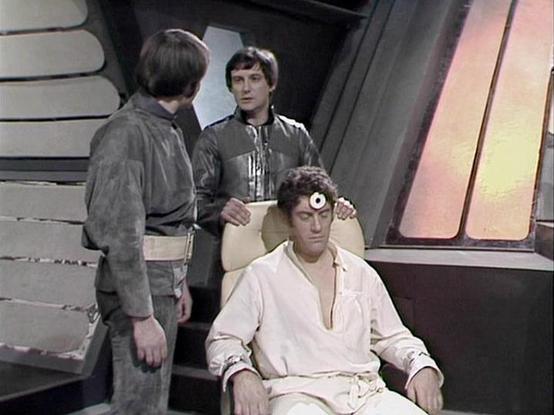 Claude Sonnet 4 describes the image as: "This appears to be a scene from the British science fiction television series "Blake's 7" (1978-1981). The setting shows the interior of what looks like a futuristic spacecraft or space station, with characteristic curved walls and metallic surfaces typical of the show's production design.

In the scene, there are three characters present. One person is seated in what appears to be a chair or command position, wearing light-colored clothing and having so…