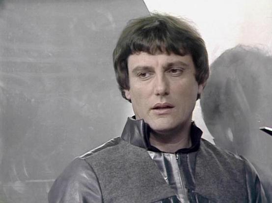 Claude Haiku 4.5 describes the image as: "# Image Description

This is a portrait photograph of an actor in what appears to be a science fiction production setting. The image shows a man with distinctive dark hair styled with bangs, wearing a gray and dark colored jacket or tunic typical of 1970s-80s television costume design. The lighting and composition suggest this is a promotional or production still from a science fiction television series.

The background is minimal and neutral, with gray…