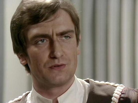 Claude Sonnet 4 describes the image as: "This image shows a man in what appears to be a military or formal uniform with decorative braiding or cord details on the shoulders. He has brown hair styled in a 1970s fashion and is looking directly at the camera with an intense expression. The background appears to be indoors with light-colored walls or pillars, suggesting this may be from a television production or film from that era. The costume and styling are consistent with period drama productio…