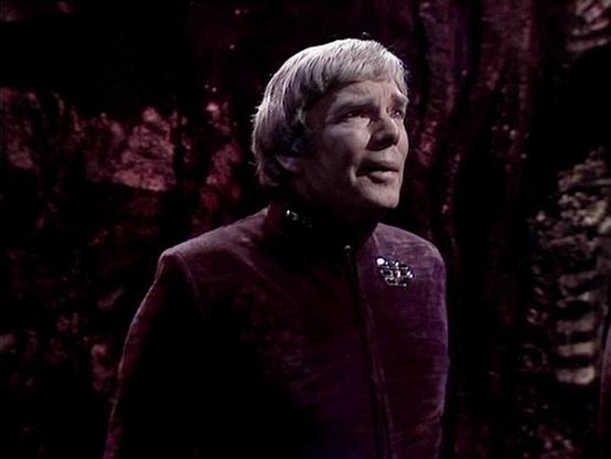 Claude Sonnet 4.5 describes the image as: "This image captures a dramatic moment from what appears to be a classical stage production or filmed theatrical performance. The actor is shown in a contemplative pose, gazing upward against a dark, textured background that suggests a cave or rocky environment. He wears a rich burgundy or wine-colored velvet tunic with an ornate brooch at the collar, costume choices that evoke medieval or Renaissance period drama.

The lighting creates a theatrical chi…