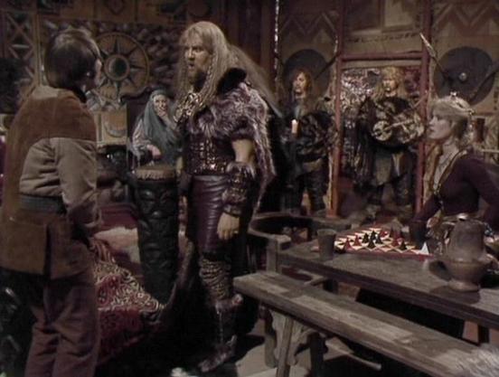 Claude Sonnet 4 describes the image as: "This appears to be a scene from the British science fiction television series "Blake's 7," set in what looks like a tavern or underground meeting place. The setting has a rustic, medieval-like atmosphere with wooden tables, stone walls, and various props scattered around including what appears to be a chess set on the table. The lighting is dim and atmospheric, typical of the show's distinctive visual style from the late 1970s/early 1980s.

The scene sho…
