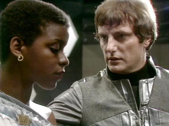 Claude Sonnet 4 describes the image as: "This image appears to be from a British science fiction television production, likely from the 1970s or 1980s based on the visual style and costume design. The scene shows two characters in what appears to be a futuristic or space-based setting. One character is wearing a distinctive metallic or silver-colored outfit with a high collar and geometric design elements typical of sci-fi costume design from that era. The other character is wearing what appear…