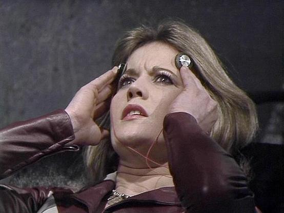 Claude Haiku 4.5 describes the image as: "# Image Description

This image shows a woman in what appears to be a scene from a science fiction television production, likely from the 1970s based on the visual style and color grading. She is wearing a burgundy or maroon-colored outfit and has styled blonde hair typical of that era. Her expression conveys distress or concern, with her hand raised to her face in a gesture suggesting worry or emotional turmoil.

The setting appears to be an interior s…