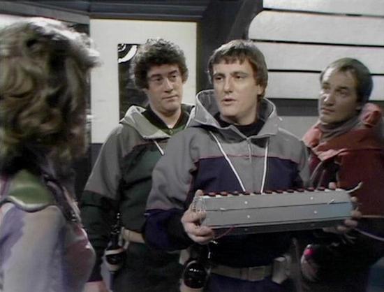 Claude Haiku 4.5 describes the image as: "# Scene from Blake's 7

This image captures a tense moment from the science fiction series *Blake's 7*, showing four cast members in what appears to be an interior spacecraft or space station setting. The scene takes place in a utilitarian corridor or control room, identifiable by the metallic walls and industrial design typical of the show's aesthetic.

The four actors are engaged in what seems to be a dramatic confrontation or discussion. One cast mem…