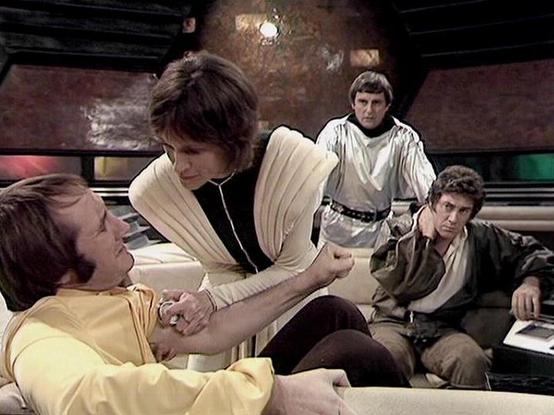 Claude Sonnet 4 describes the image as: "This appears to be a scene from the British science fiction television series "Blake's 7," which aired from 1978-1981. The image shows what looks like a medical or care scene taking place aboard the spaceship Liberator. The setting features the show's characteristic futuristic interior design with curved walls and distinctive lighting. The scene appears to be taking place in one of the ship's medical or living areas, with the crew gathered around someone…