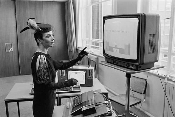 British actress Jacqueline Pearce inspects a Texas Instruments TI-99-4 computer with Speech Synthesiser enabling it to talk, play chess and table tennis, as well as keep accounts, United Kingdom, 10th April 1980. (Photo by Geoff Bruce/Central Press/Hulton Archive/Getty Images)

In this black-and-white photograph, a woman in striking avant-garde fashion stands before a vintage computer setup, likely in the early 1980s. She wears a dark, long-sleeved dress with sequined "cold-shoulder" cutouts, l…