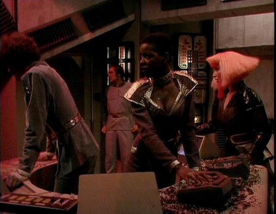 Claude Sonnet 4.5 describes the image as: "This image appears to be from a science fiction television production, showing a scene set in what looks like a spacecraft or futuristic facility interior. The setting features technical panels and equipment typical of sci-fi set design from the late 1970s or early 1980s.

The scene depicts four individuals in costume around what appears to be a control station or work area. The costumes are characteristic of space opera productions of that era, featur…