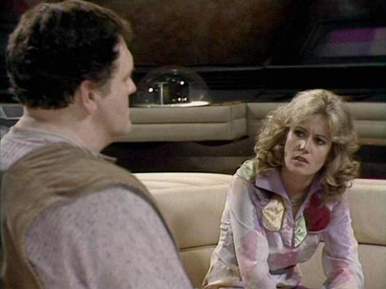 Claude Sonnet 3.7 describes the image as: "The image shows a scene from what appears to be a television program, likely from the 1980s based on the visual style and fashion. Two people are seated in what looks like the interior of a spaceship or futuristic vehicle with cream-colored seating.

On the right is a person with curly blonde hair wearing a distinctive patterned blouse with pastel colors including pink, yellow, and lavender. This character appears to be engaged in serious conversation.…