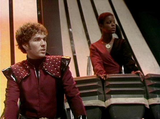 Claude Haiku 4.5 describes the image as: "# Scene Description

This image captures a scene from what appears to be a science fiction television production, set in a futuristic spacecraft interior. The setting features distinctive angular architectural elements with metallic striping on the walls, creating a sleek, modernist aesthetic typical of 1980s sci-fi design.

The character on the left wears an elaborate burgundy uniform with a distinctive studded shoulder cape, suggesting a high-ranking …