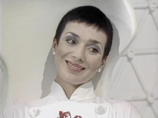 Claude Sonnet 4 describes the image as: "I can see someone in what appears to be a futuristic white costume with distinctive circular elements or badges on the chest. The person has very short, dark hair in a distinctive pixie cut style and is wearing small earrings. The setting appears to be from a science fiction production, with white, curved architectural elements visible in the background that give it a sleek, futuristic appearance. The costume design and overall aesthetic suggests this is…