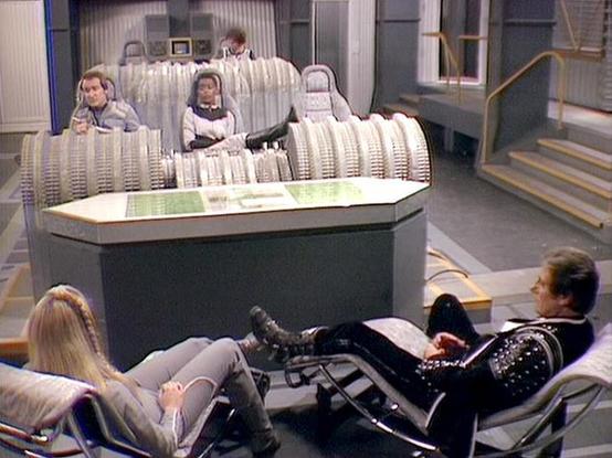 Claude Sonnet 4 describes the image as: "This appears to be a scene from the British science fiction television series "Blake's 7." The setting shows a futuristic spacecraft interior with metallic, ribbed walls and angular architecture typical of the show's distinctive production design. Several crew members are positioned around what appears to be the ship's command center or medical bay area. The scene has the characteristic low-budget but imaginative aesthetic that "Blake's 7" was known for,…