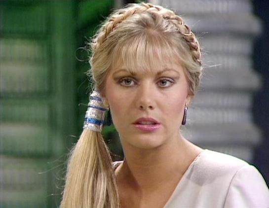 Claude Haiku 4.5 describes the image as: "# Image Description

This image shows a woman with blonde hair styled in a distinctive 1980s fashion, featuring a braided crown detail and a long ponytail with blue and white beaded hair accessories. She has fair complexion, defined makeup with pink lipstick, and is wearing what appears to be a light-colored top or dress.

The setting appears to be an indoor location with a blurred background showing green and grey tones, suggesting an institutional or …