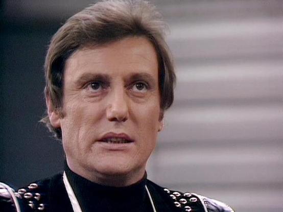 Claude Haiku 4.5 describes the image as: "# Image Description

This image shows a man in character wearing a distinctive black outfit adorned with metallic studs and decorative elements. The styling, including the shoulder armor and embellished costume, suggests a science fiction production from the 1970s or 1980s. The character has dark hair styled in a period-appropriate fashion and displays an intense, serious expression.

The background features neutral gray tones with horizontal lines, con…