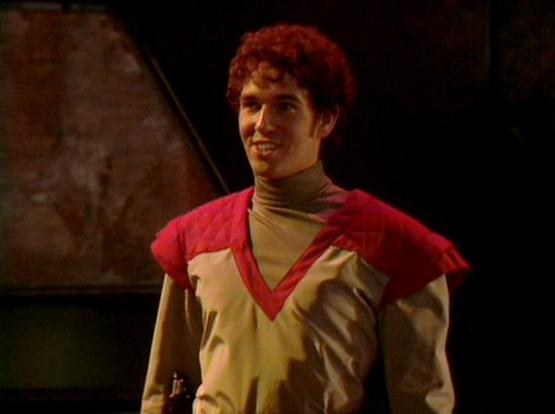 Claude Haiku 4.5 describes the image as: "# Image Description

This image shows an actor in a theatrical or television production setting against a dark, minimalist backdrop. The performer is wearing a distinctive costume consisting of a tan or beige long-sleeved turtleneck paired with a red sleeveless tunic or vest layered over it. The character has reddish curly hair styled in a voluminous manner typical of 1980s-90s television production.

The lighting is warm and theatrical, creating an int…