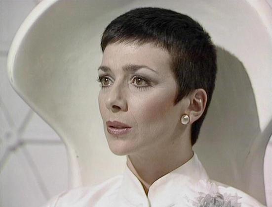 Claude Haiku 4.5 describes the image as: "# Image Description

This image shows a woman in profile, positioned against a minimalist, curved white background that creates a stark, clinical aesthetic. She wears a white high-necked garment with delicate embroidered details on the chest, contributing to a refined, formal appearance.

Her styling is distinctive: she has short, dark hair cut close to the head, and her makeup is precise and dramatic, featuring prominent eye makeup that emphasizes her …