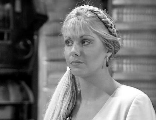 Claude Sonnet 4 describes the image as: "This black and white image shows a woman with blonde hair styled in an elaborate braided updo with bangs. She's wearing what appears to be a white or light-colored garment and dangling earrings. The woman is photographed in profile, looking off to the side with a thoughtful or contemplative expression. The setting appears to be indoors with some brick or stone architecture visible in the blurred background. The lighting and composition suggest this is fr…