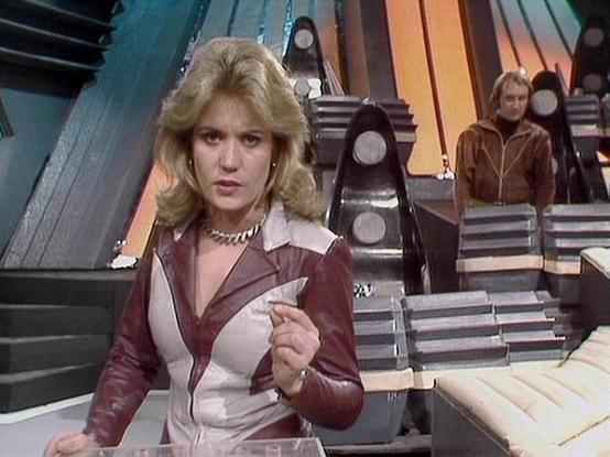 Claude Haiku 4.5 describes the image as: "This image appears to be from the classic science fiction television series "Blake's 7," showing a futuristic spacecraft interior setting. The scene features a woman in the foreground wearing a distinctive maroon and white outfit with a collar detail, holding what appears to be a document or tablet. She has blonde, voluminous 1970s-style hair and is positioned at what looks like a control station or desk.

Behind her, the set displays the characteristic…