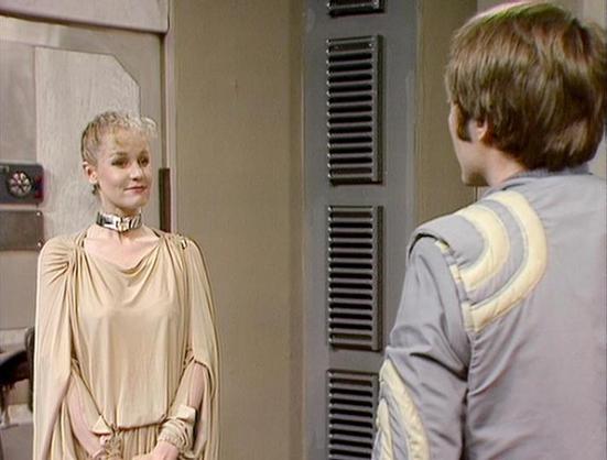 Claude Haiku 4.5 describes the image as: "# Scene Description

This image appears to be from a science fiction television production, showing two characters in what looks like a spaceship or futuristic facility corridor. The setting features distinctive metallic ventilation panels and neutral-toned walls typical of 1970s-80s sci-fi aesthetics.

On the left stands a character wearing an elegant flowing cream-colored robe or gown with draped fabric and a decorative collar piece. The styling inclu…