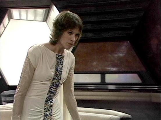 Claude Sonnet 3.7 describes the image as: "The image appears to be a still from a television series, showing a person in a distinctive white outfit with a floral or patterned panel down the front. They're standing in what looks like a futuristic or sci-fi setting, with angular walls and panels visible in the background. The interior space has a geometric design with light panels contrasting against darker structural elements.

The costume has a 1970s-80s sci-fi aesthetic, with a cream-colored f…