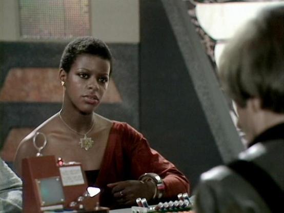 Claude Sonnet 4.5 describes the image as: "This image shows a scene from what appears to be a science fiction production, likely from the 1980s based on the aesthetic and film quality. The setting features a futuristic interior with industrial-style architecture, including angular metal beams and muted earth-toned wall panels visible in the background.

The actress Josette Simon is featured prominently in the frame, wearing a rust-colored or burgundy uniform with metallic shoulder detailing and…