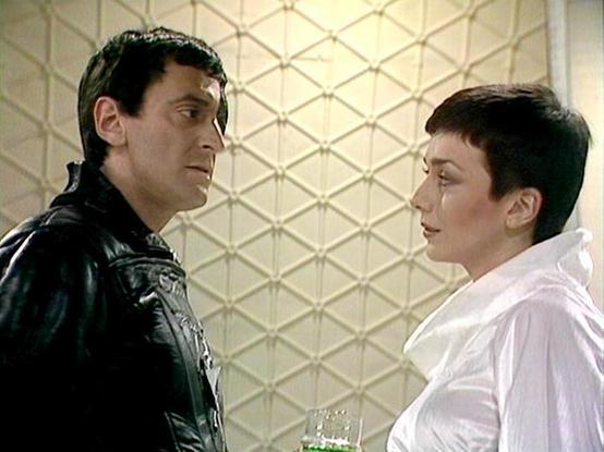 Claude Sonnet 4 describes the image as: "This image shows two actors in what appears to be a science fiction television production, likely from the late 1970s or early 1980s based on the styling and production values. The scene is set in a futuristic interior with distinctive geometric wall paneling featuring a diamond or lattice pattern in light colors. One actor wears an elaborate black leather or vinyl costume with ornate detailing, while the other is dressed in a flowing white garment. The …