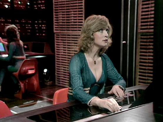 Claude Sonnet 3.7 describes the image as: "This image appears to be from a vintage television production, likely from the 1970s or early 1980s based on the styling and production quality. The scene shows a person with blonde, curly hair wearing a teal green outfit with a v-neck design and a small pendant necklace. They appear to be working at some kind of control desk or console, possibly in a broadcast studio or communications setting, as suggested by the equipment in front of them and the win…