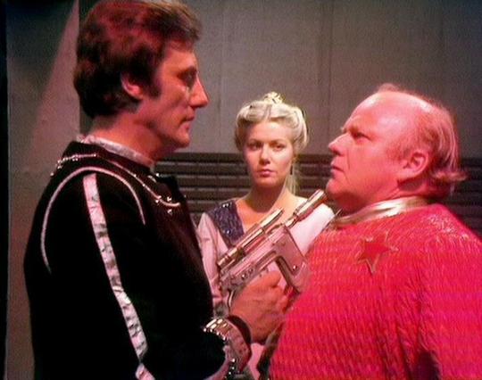 Claude Sonnet 3.7 describes the image as: "This image appears to be from a science fiction TV show, likely from the late 1970s or early 1980s based on the production style and costuming. The scene shows three performers in what looks like a confrontation on a spaceship or futuristic set.

On the left is a person wearing a black costume with silver detailing that has a military or authoritarian style. In the center stands someone with blonde hair styled in an elaborate updo, wearing what appears…