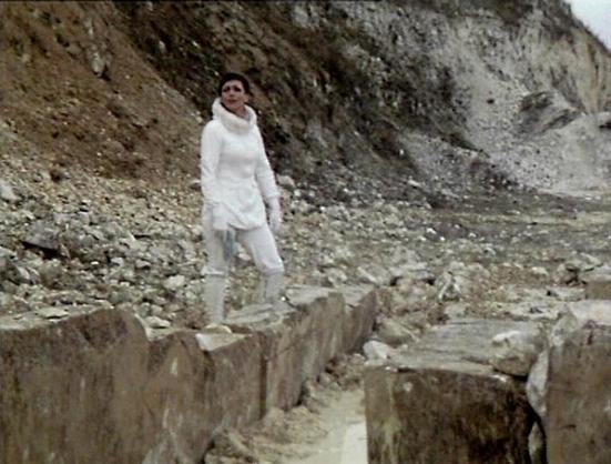 Claude Sonnet 4 describes the image as: "This image shows a scene from what appears to be a science fiction production, set in a rocky, quarry-like landscape with chalk or limestone cliffs in the background. The terrain is rough and barren, with scattered rocks and debris creating an alien or otherworldly atmosphere typical of classic British sci-fi productions from the 1970s-80s.

A figure in a flowing white costume stands prominently in the scene, positioned on elevated rocky ground. The whit…