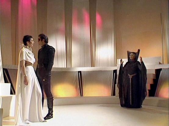 Claude Sonnet 4 describes the image as: "This image appears to be from a science fiction television production, likely from the 1970s or early 1980s based on the aesthetic and production values. The scene takes place in a futuristic, minimalist set with sleek metallic walls, geometric architectural elements, and atmospheric lighting in warm tones of gold and pink.

The setting appears to be some kind of ceremonial or formal space, possibly a throne room or council chamber, with its clean lines …