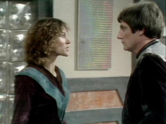 Claude Sonnet 3.7 describes the image as: "This image appears to be from a vintage television program, likely from the 1970s or 1980s based on the visual style and production quality. The scene shows two people facing each other in what seems to be an interior setting with distinctive architectural elements, including a textured wall panel and some glass brick elements visible on the left side.
The person on the left is wearing a dark outfit with a teal or turquoise collar detail, while the pe…