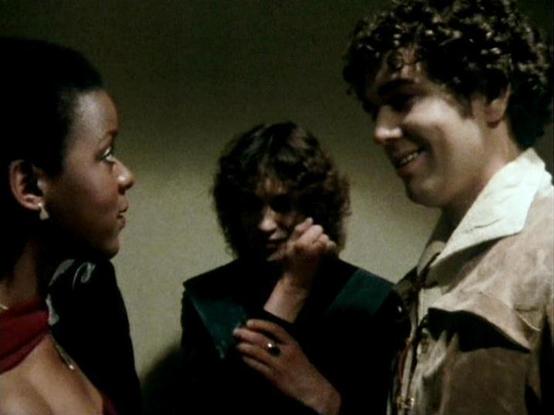 Claude Sonnet 4.5 describes the image as: "This image captures a tense dramatic moment featuring three actors in what appears to be a scene from a television production, likely from the late 1970s or early 1980s based on the film quality and styling. The setting is intimate and claustrophobic, with muted olive-green walls creating an oppressive atmosphere.

On the left stands Josette Simon, wearing a striking red and black costume with shoulder detailing, her gaze directed intensely at the othe…