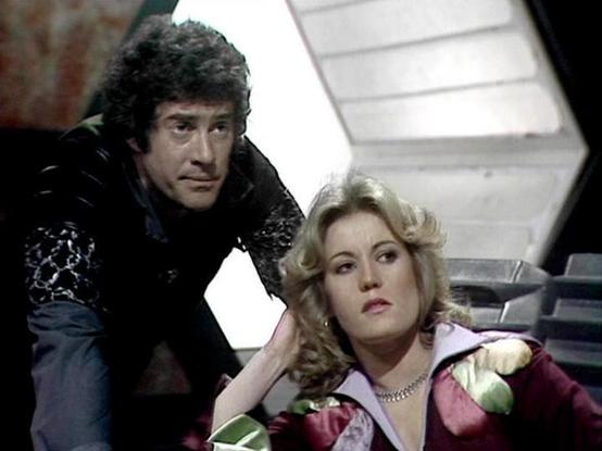 Claude Sonnet 4 describes the image as: "This image appears to be from a classic science fiction television production, likely from the late 1970s or early 1980s based on the visual style and production values. The setting appears to be the interior of a spacecraft or futuristic facility, with characteristic angular white paneling and technological elements visible in the background.

The scene shows two characters in what appears to be a dramatic moment. One figure wears dark leather or simila…