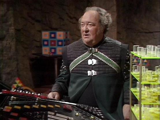 Claude Sonnet 3.7 describes the image as: "This image shows a scene from what appears to be a science fiction television production, likely from the classic BBC series "Doctor Who." The setting features a futuristic control room with a stone-like wall background. 

The person in the image is wearing an elaborate costume consisting of a dark quilted or textured tunic with green panels and decorative white fastenings. They appear to be standing at some sort of control panel with buttons and color…