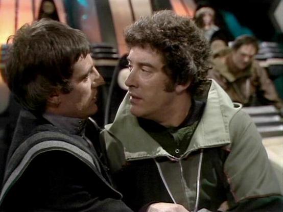 Claude Sonnet 4 describes the image as: "I can see this appears to be a scene from what looks like a science fiction television production, likely from the late 1970s or early 1980s based on the visual style and production quality. The image shows two men in close conversation - one wearing dark clothing and the other in lighter colored outerwear with what appears to be a hooded jacket or vest. The setting appears to be some kind of futuristic or institutional environment with other people visi…