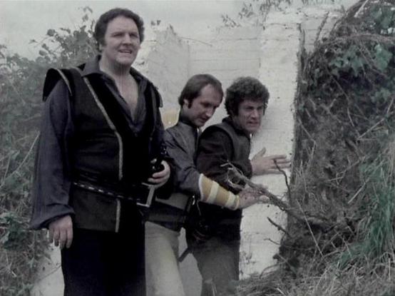 Claude Sonnet 4.5 describes the image as: "This image appears to be from the British science fiction television series "Blake's 7," which aired from 1978 to 1981. The scene shows three actors in costume on what appears to be an outdoor set with white walls and overgrown vegetation.

The actors are dressed in distinctive period costumes typical of the show's aesthetic. The figure on the left wears a black outfit with striking metallic trim or piping, while the two figures on the right are dresse…